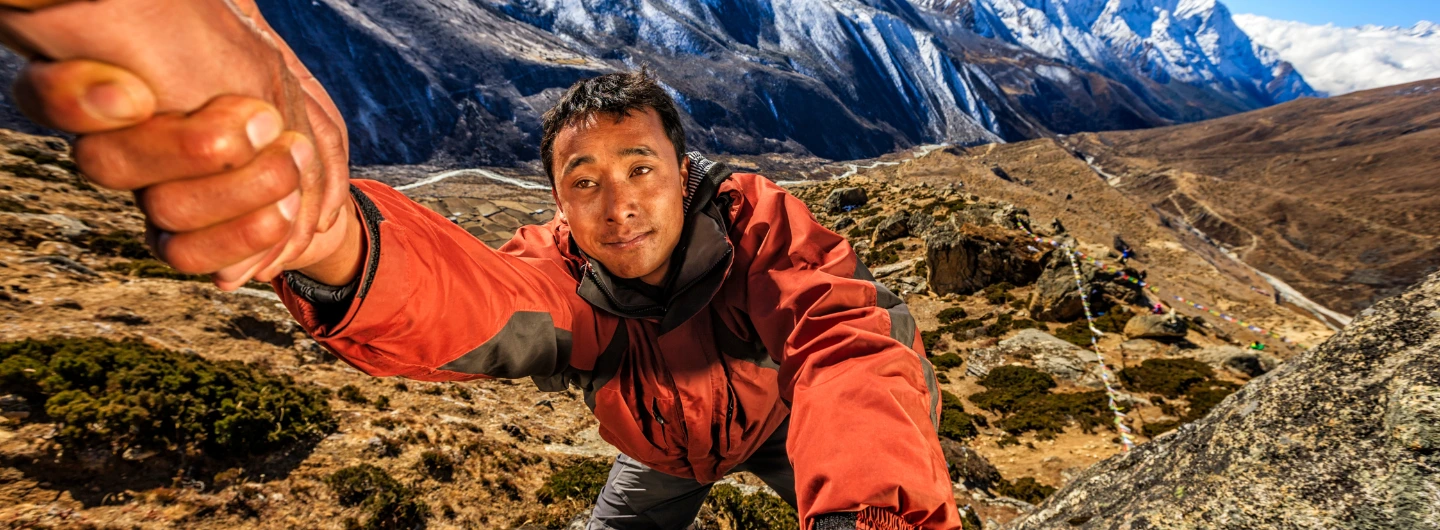 Climber in red jacket reaching for a helping hand on rocky slope, symbolizing how Sherpas help climbers conquer tough mountain terrain, with snowy peaks and valley in background