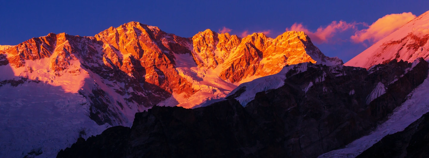 Snow-covered peaks glowing at sunset along the Kanchenjunga trekking route, with vibrant orange-pink light, dark shadows, and a deep blue sky