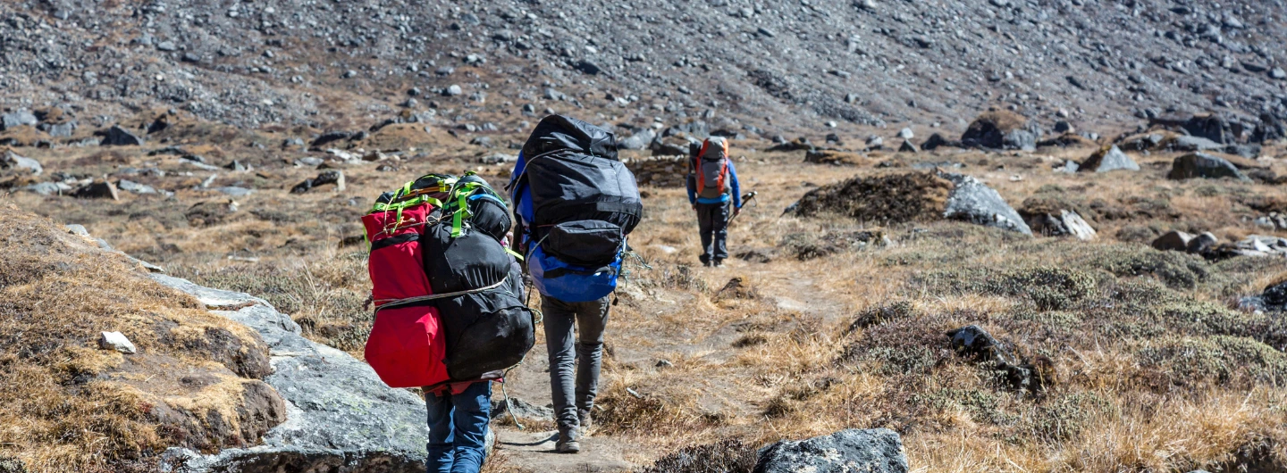 Three hikers with large backpacks trekking a rocky mountain trail, illustrating why Sherpas are helpful in climbing expeditions for navigating tough terrain