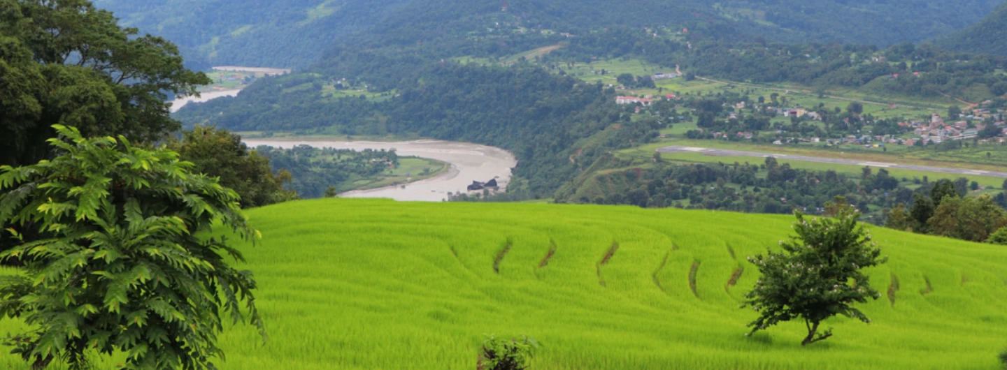 Green terraced fields with trees overlooking a winding river and small town on the Kanchenjunga Trek route
